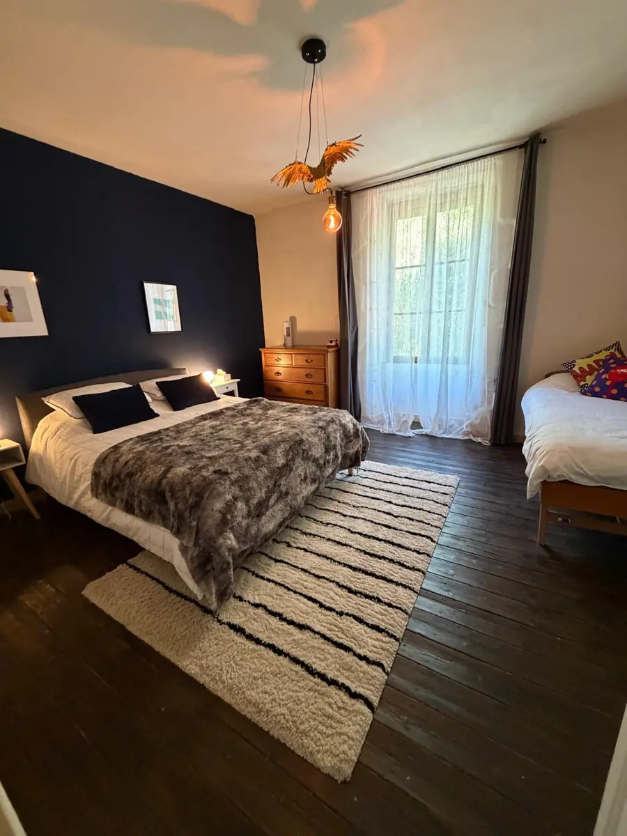 Bedroom One with the lamps on: a navy feature wall behind a king bed with a faux-fur throw, a striped rug on dark floorboards, a gilded bird pendant light and a single bed by the tall window
