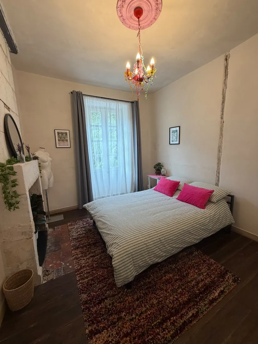 Bedroom with a double bed dressed in green striped bedding with pink cushions, an old stone fireplace, a tall window with lace curtains and a colourful chandelier below a pink ceiling rose
