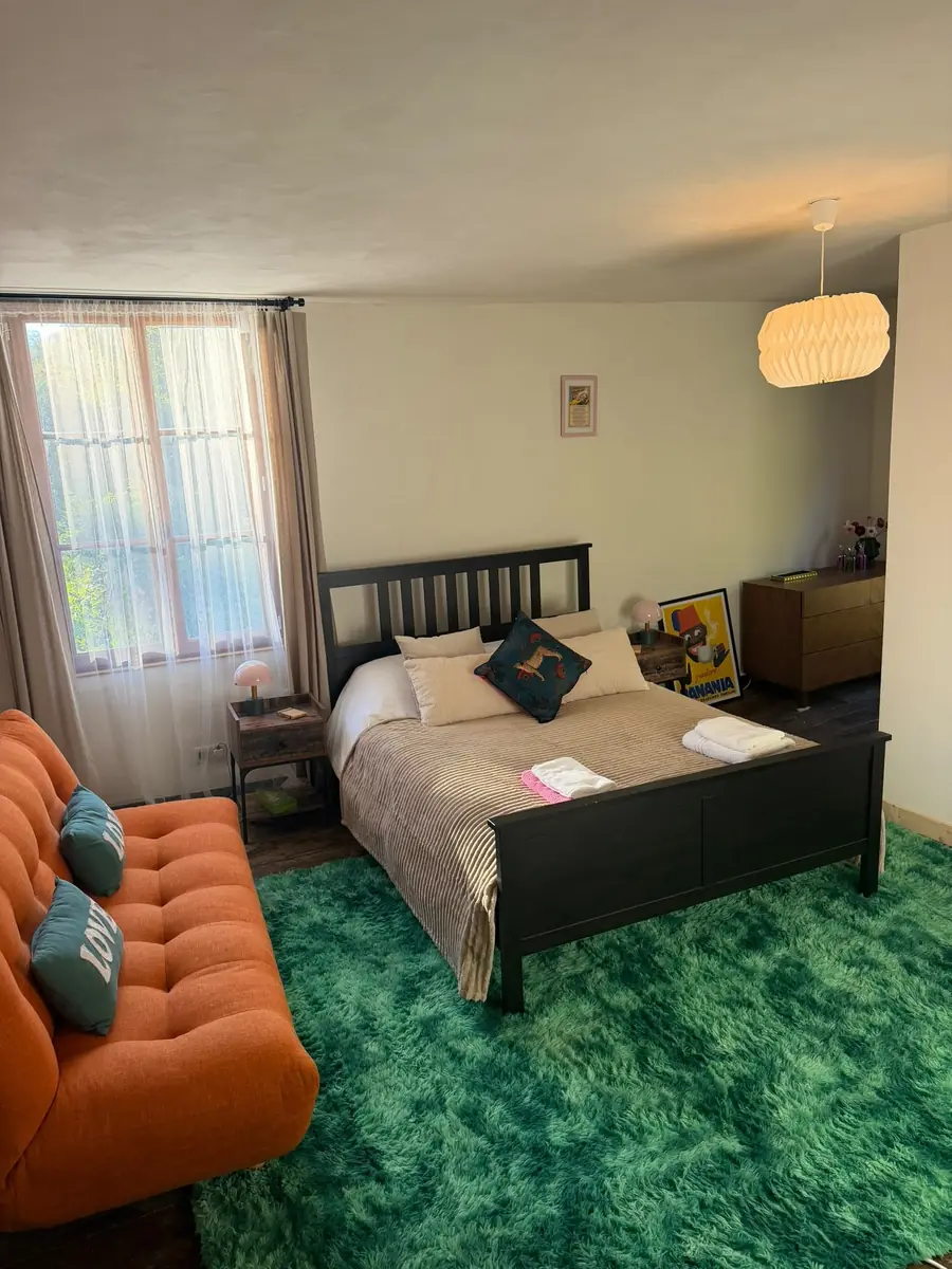 Bedroom Three: a dark wooden double bed with pale bedding, a long orange sofa on a bright green rug, a tall curtained window and a paper pendant light