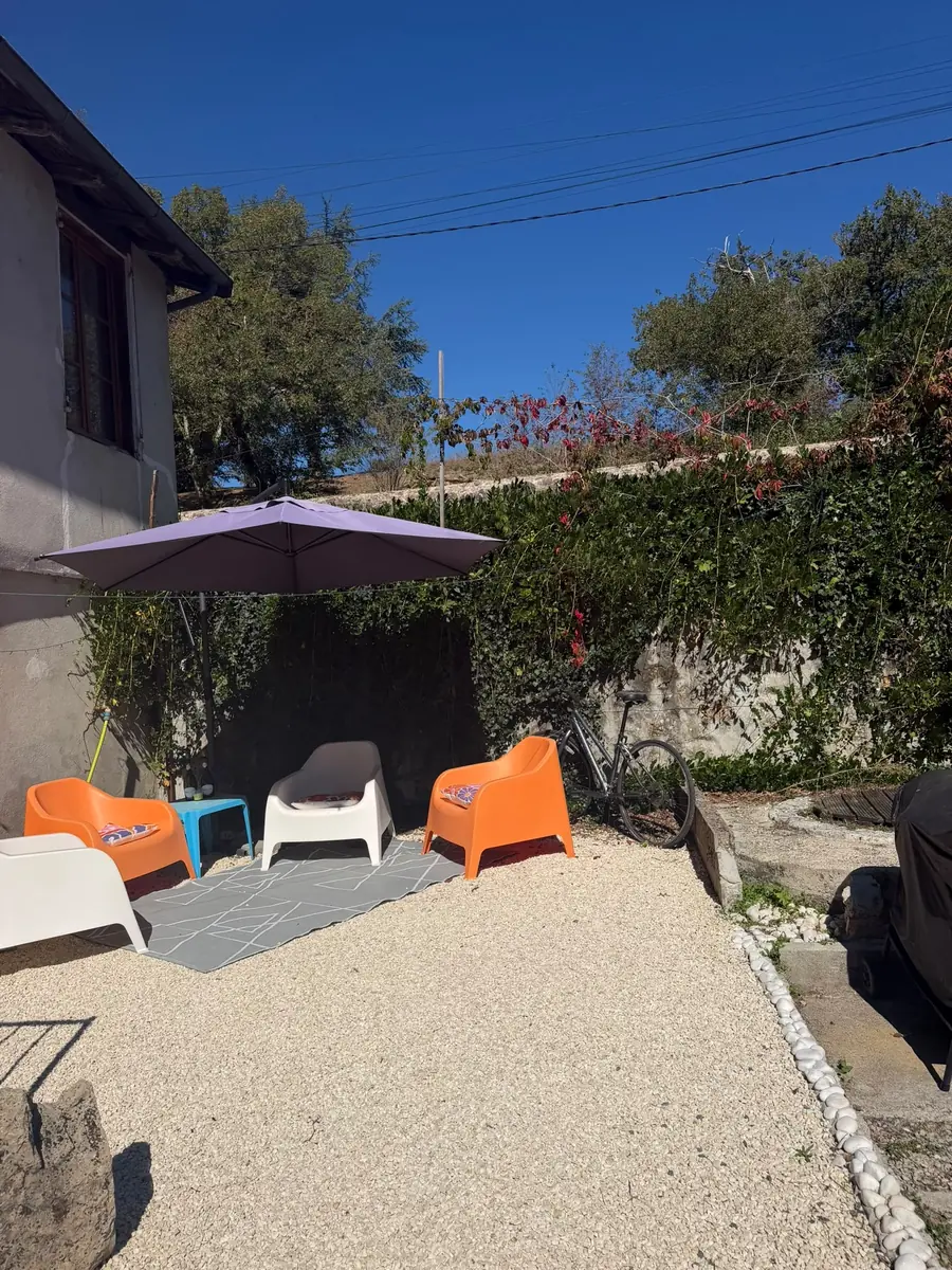 A gravel terrace beside the house with orange and white chairs on a grey rug under a purple parasol, against an ivy-covered stone wall
