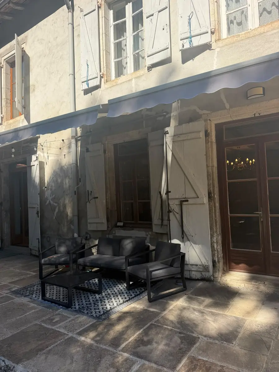 The stone-flagged patio along the back of the house: black outdoor sofas and armchairs on a patterned rug, under wind-out awnings, with the tall shutters open