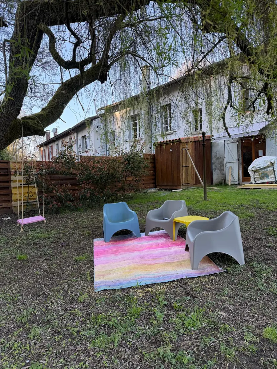 Under the willow: pale blue and grey garden chairs and a small yellow table on a pastel striped rug, a pink rope swing hanging from a branch, and the white house behind