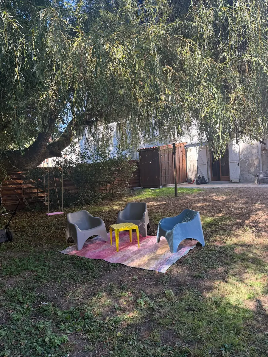 Under the willow: pale blue and grey garden chairs round a small yellow table on a pink striped rug, in dappled shade on the lawn, with the house behind