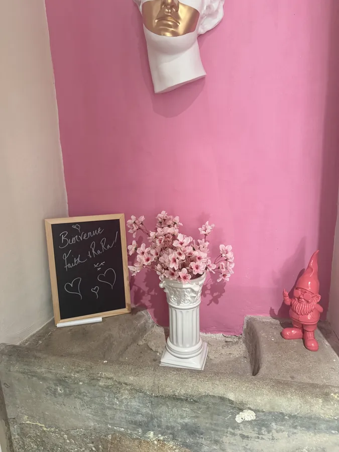 A pink-painted corner with a chalkboard reading Bienvenue, a vase of blossom on a stone plinth and a small pink ornament