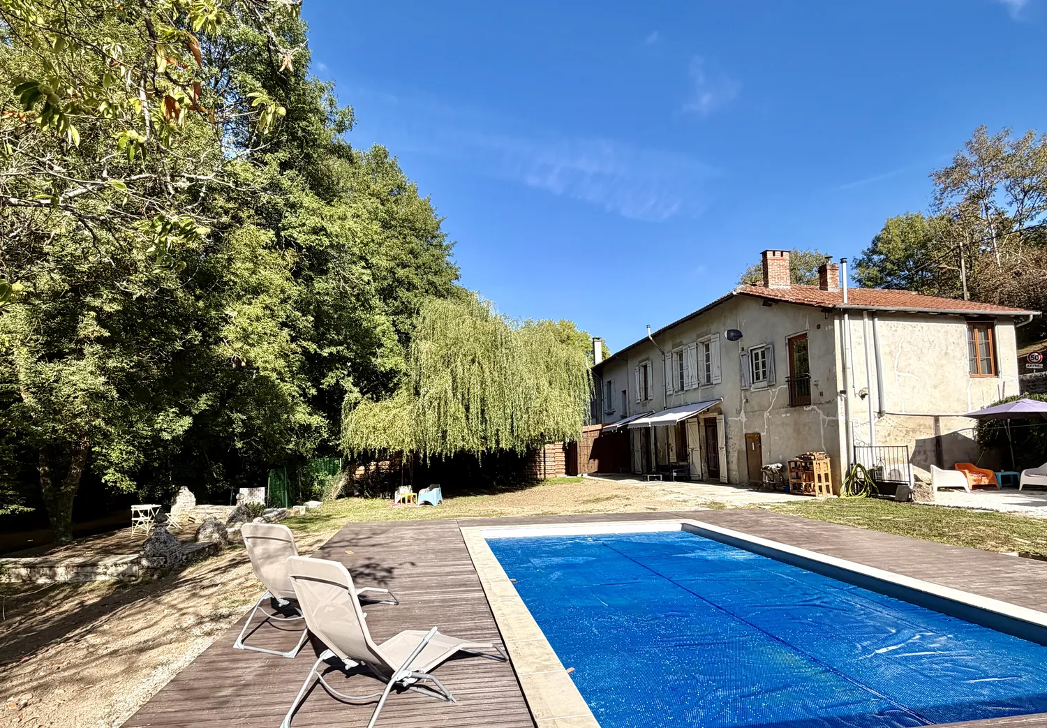 The long stone house seen from across the garden, with the swimming pool in the foreground under a blue sky
