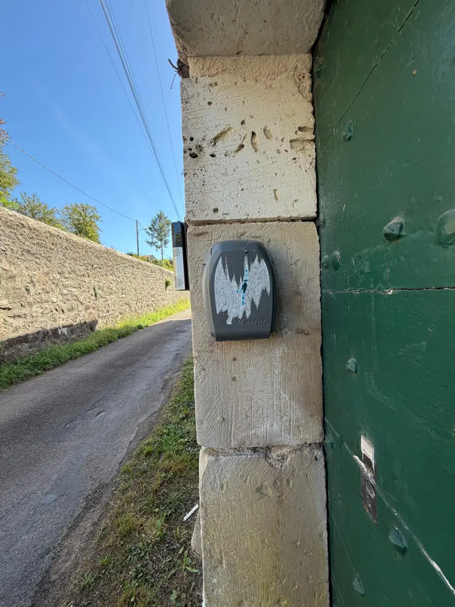 A small grey key box fixed to the pale stone frame beside the green front door, with the lane running off behind