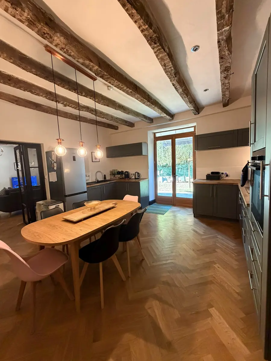 The kitchen under old timber beams: an oval wooden dining table with black and pink chairs on a herringbone floor, copper pendant lights, grey units along both sides and a glazed door to the garden at the far end