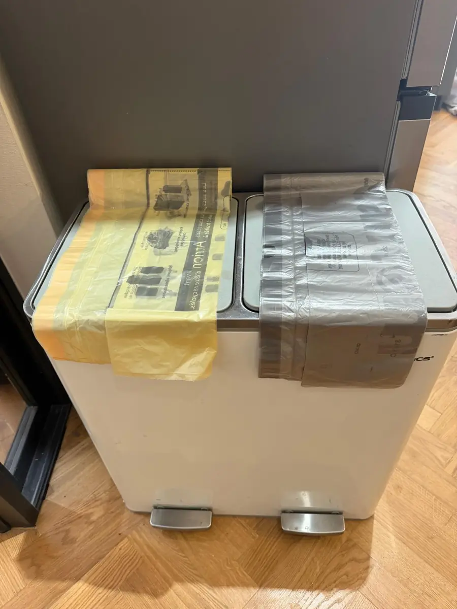 A white double pedal bin beside the fridge-freezer, one side lined with a yellow bag and the other with a grey bag