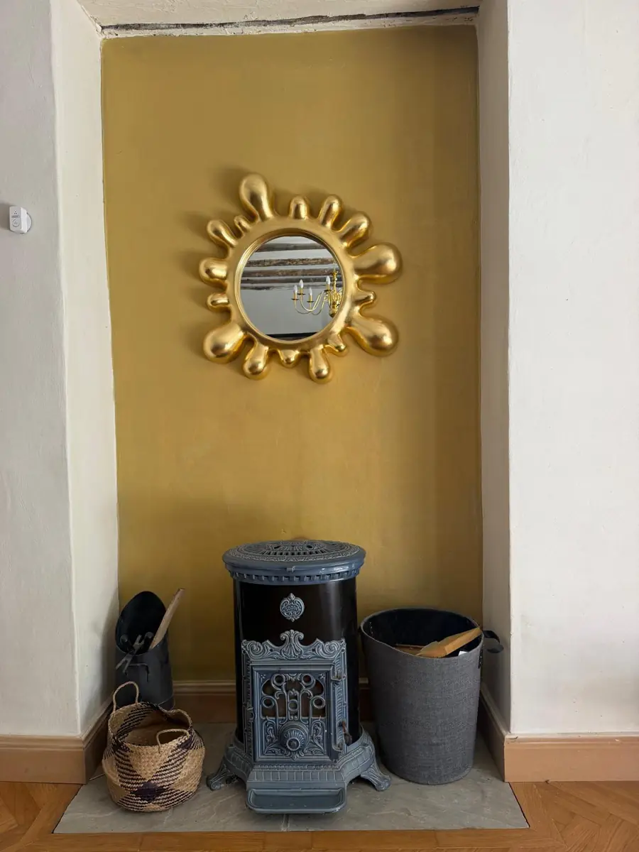The wood burner in a warm yellow alcove, under a gold sunburst mirror, with a log basket and a coal bucket either side