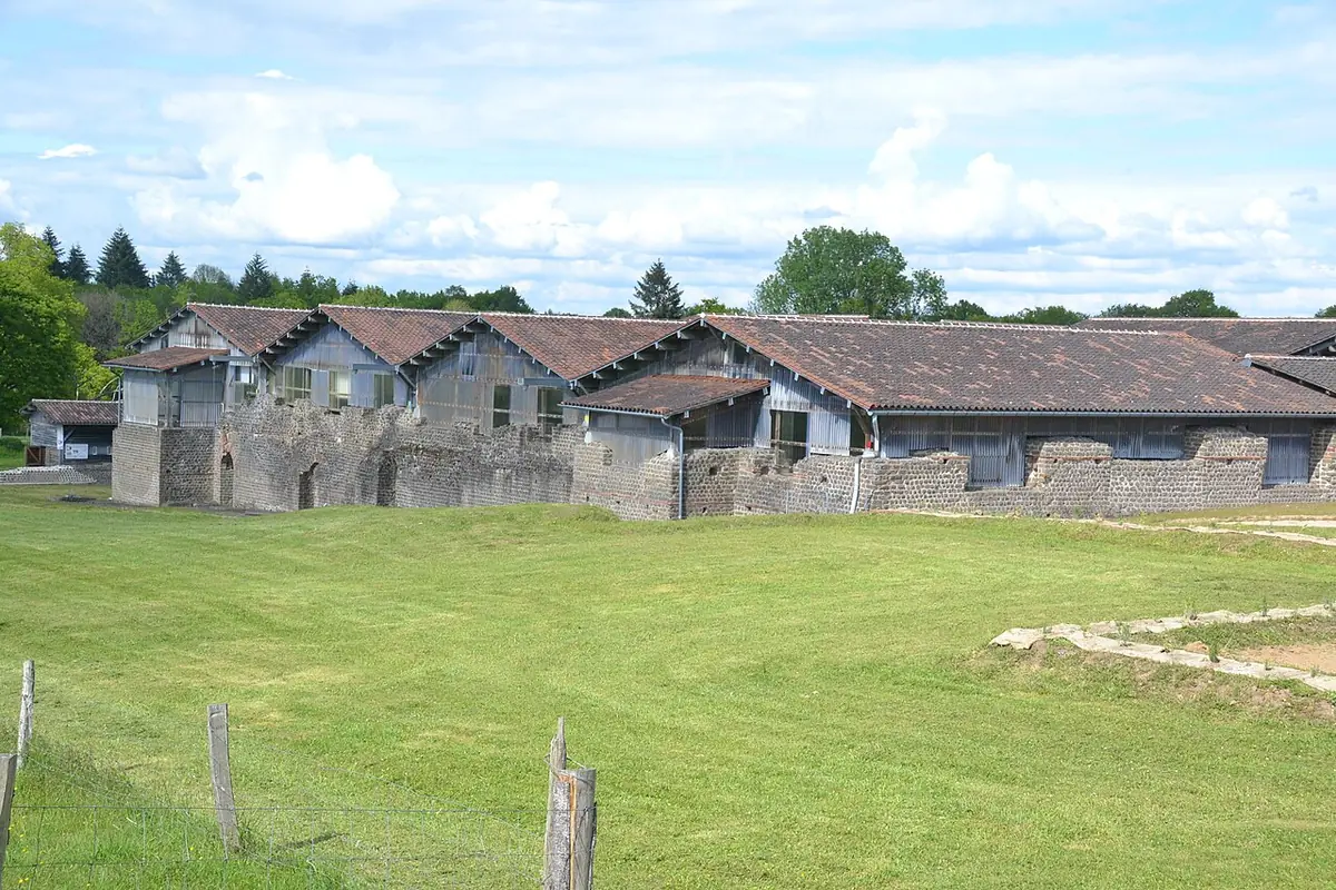 The long low stone ruins of the Roman baths at Chassenon under timber roofs, across a green field