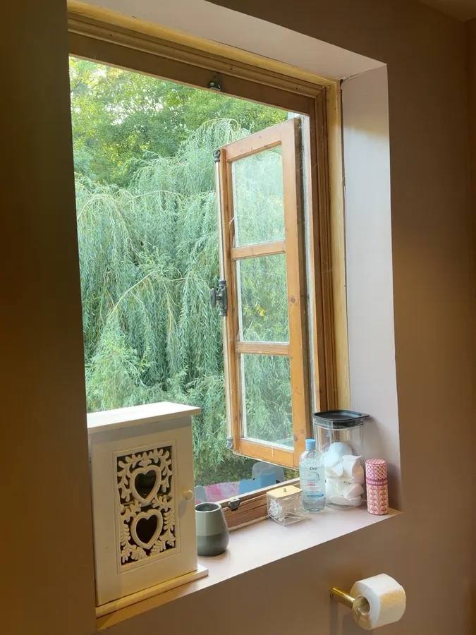 A deep-set wooden casement window standing open onto the willow tree, with jars and toiletries on the sill