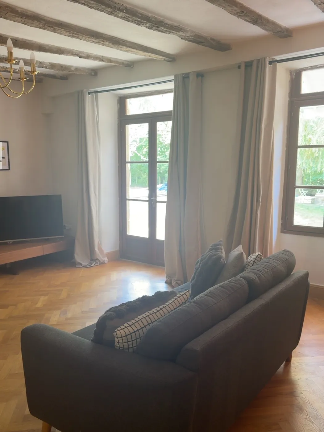 The lounge from behind the sofa: herringbone parquet floor, exposed ceiling beams, tall windows with long curtains and a television on a low unit