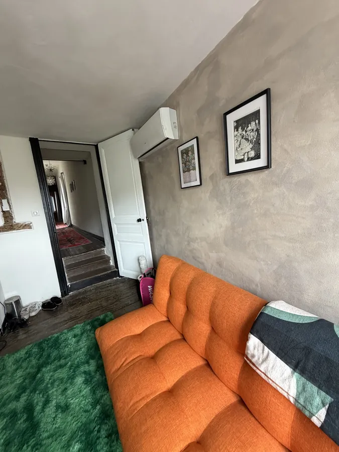 The bedroom seen from the corner, with a long orange sofa, a green rug and the door through to the hall