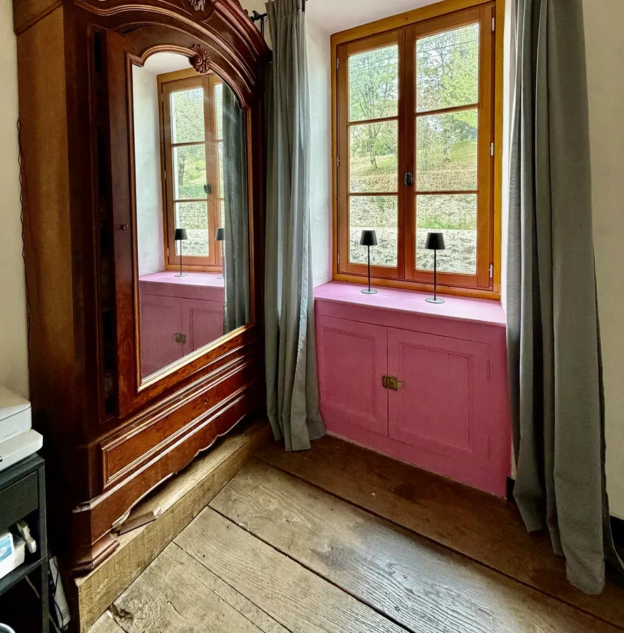 A large carved wooden armoire beside a window, with a pink-painted built-in cupboard below it