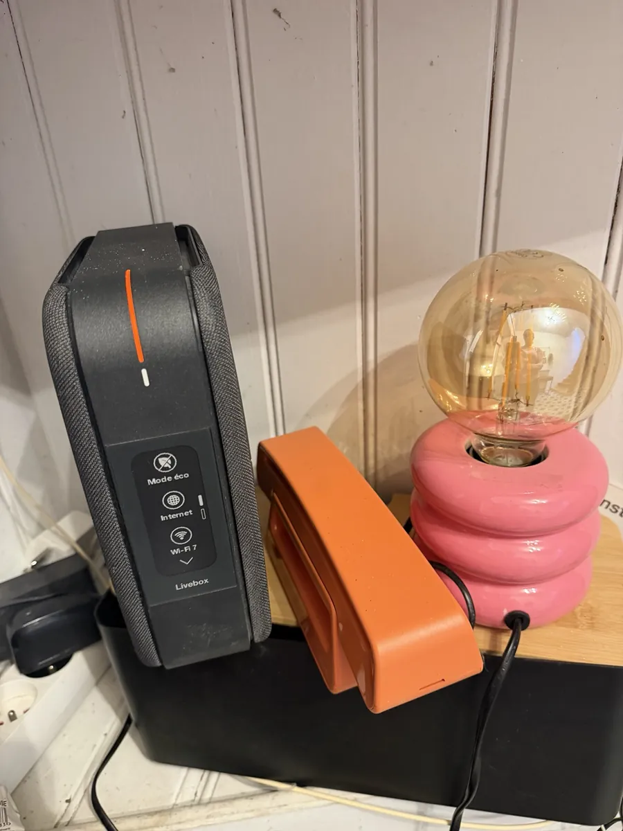 An orange and black Livebox router on a shelf beside an orange telephone handset and a small pink glass lamp