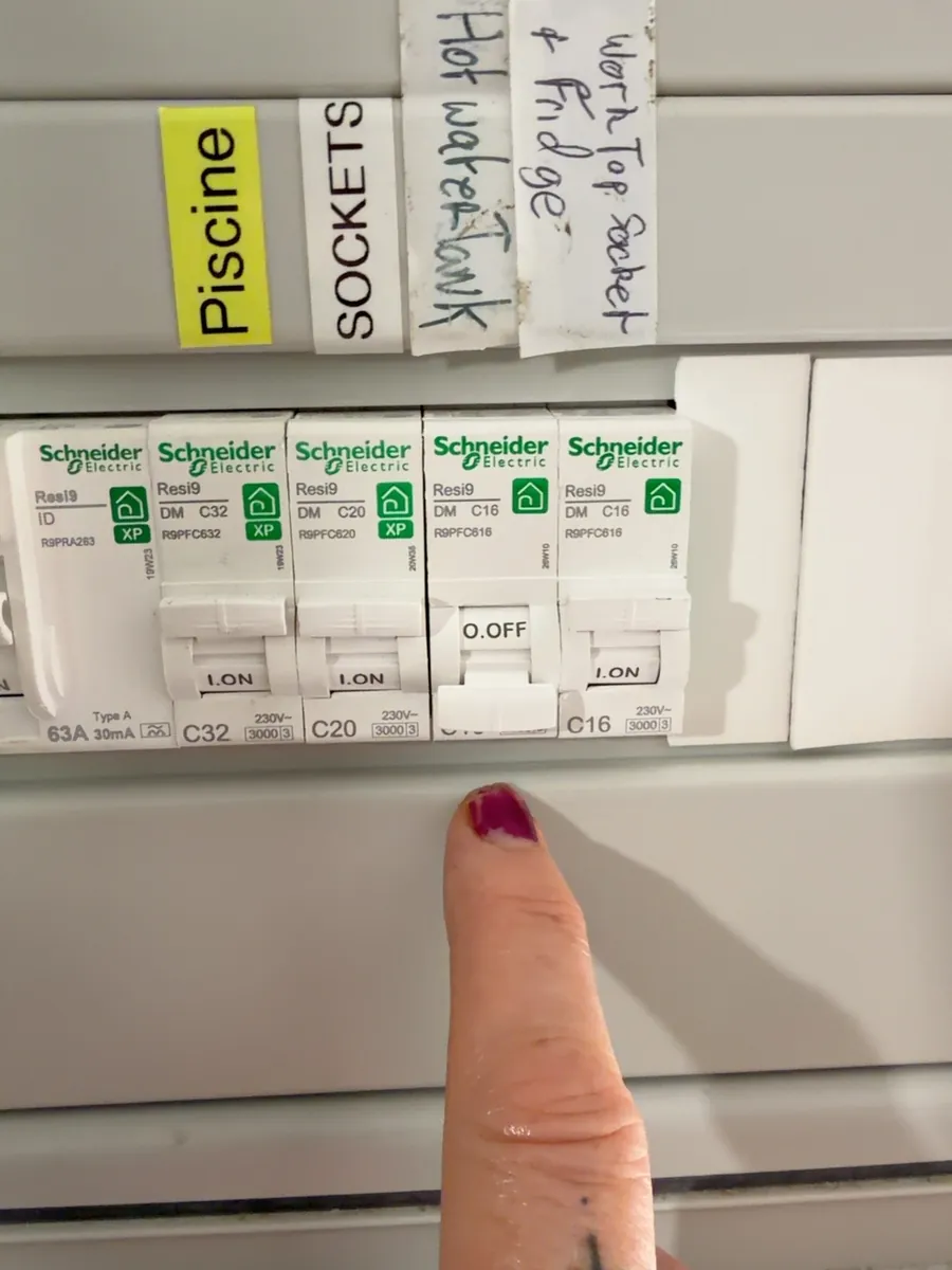 Close-up of the garage electrical board, a hand pointing to a breaker labelled hot water tank, switched off