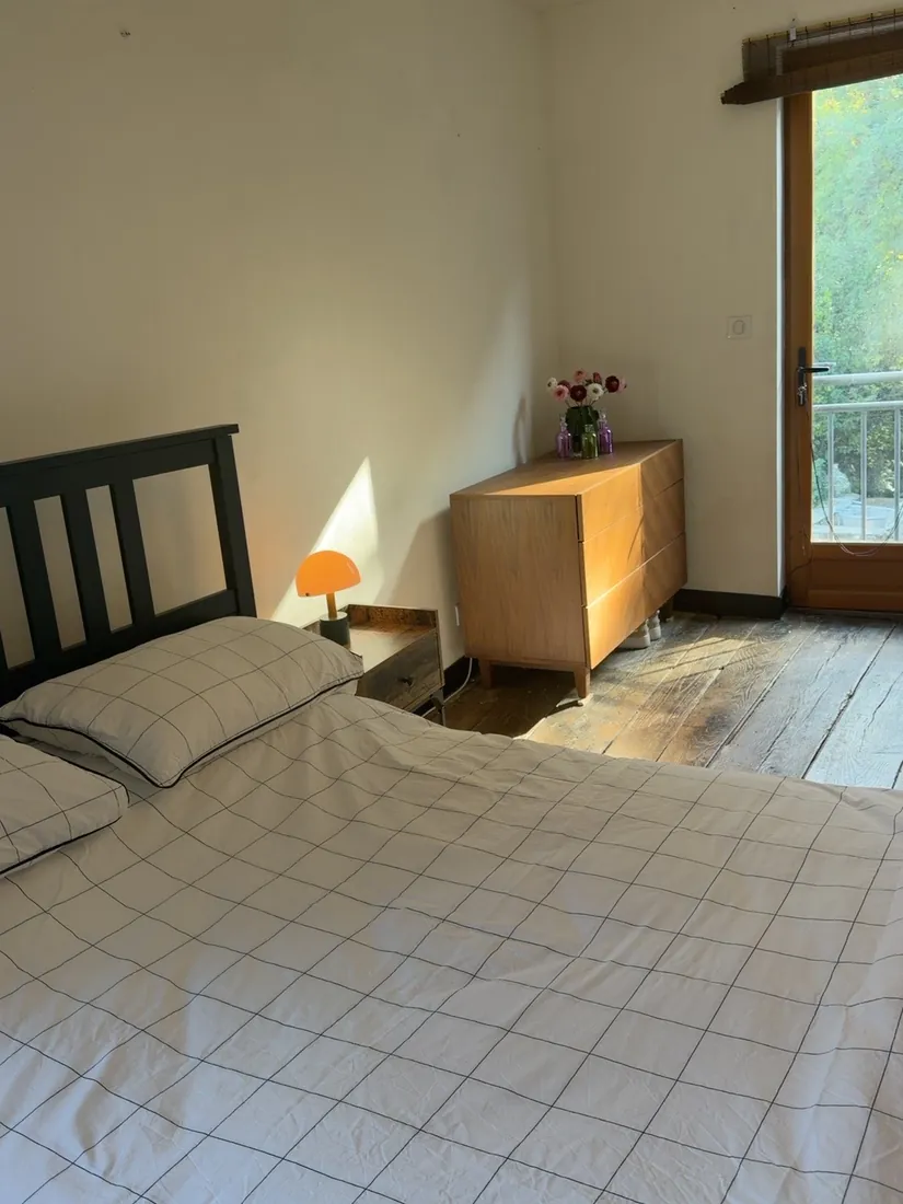 Bedroom with a dark wooden bed frame and pale bedding, a bedside lamp, a wooden chest of drawers and a glazed door onto greenery