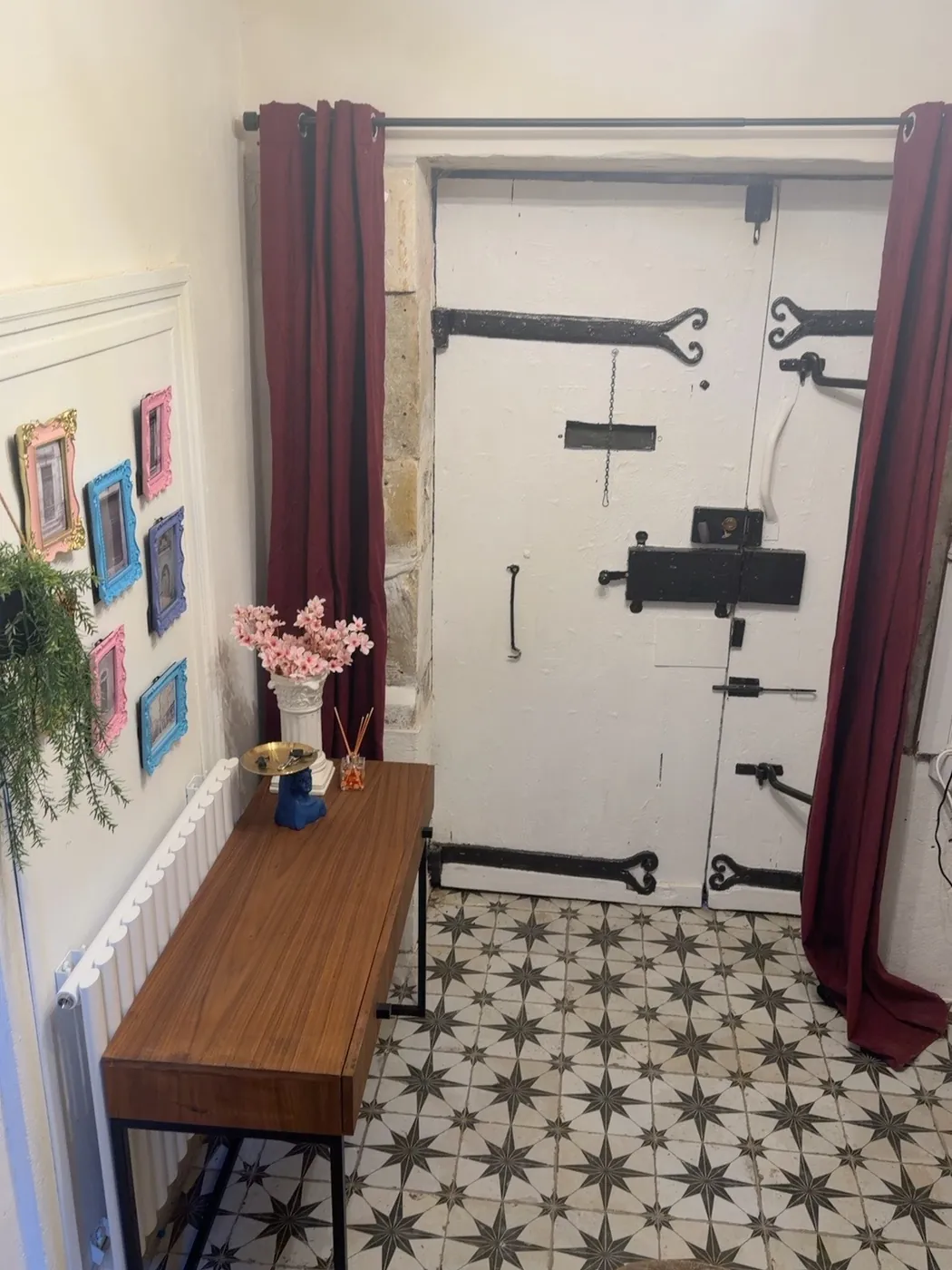The entrance hall: a patterned black and white star-tiled floor, deep red curtains framing the old white door, and a wooden bench with pink flowers