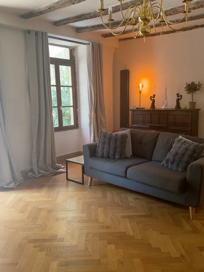 The lounge in warm evening light, with a grey sofa, a lit lamp on a sideboard and the brass chandelier overhead