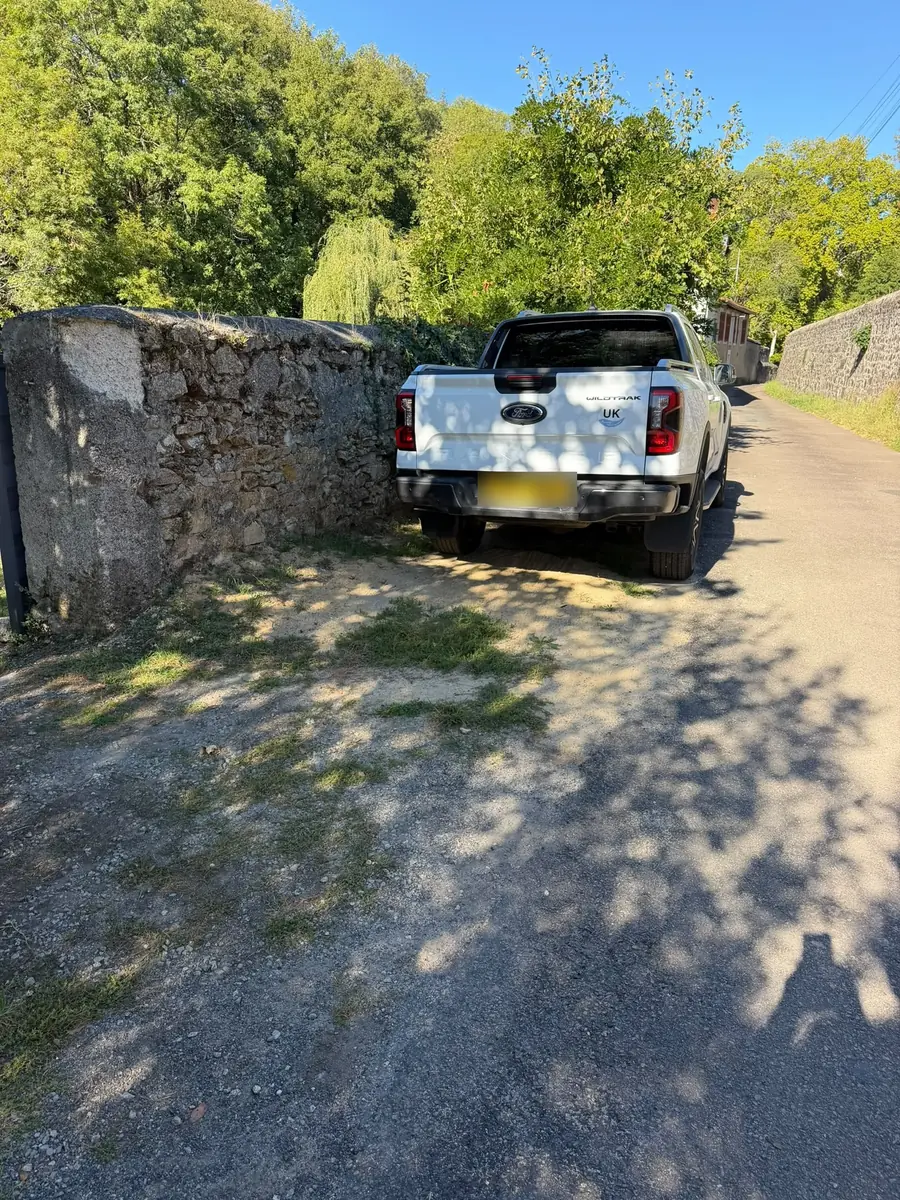 A white pickup parked on the gravel beside a high stone wall, just outside the gates, with the lane running on past it