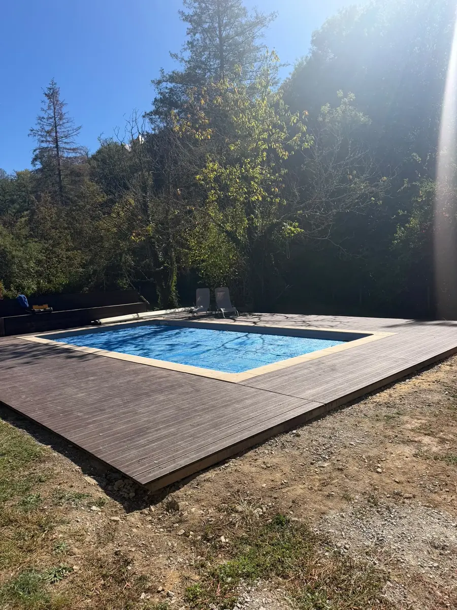The pool and its timber decking in bright sunshine, with tall trees behind
