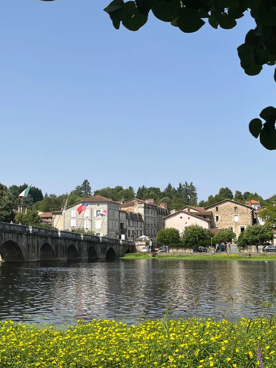 The old stone bridge across a wide, still river, with the houses of Confolens climbing the far bank under a clear sky and yellow flowers in the foreground