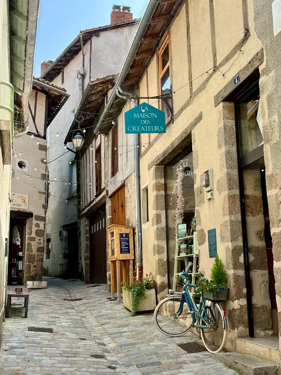 A narrow cobbled lane of stone houses, with a teal hanging sign for the Maison des Cr&eacute;ateurs, a bicycle and pots of flowers