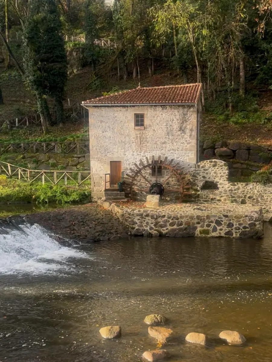 An old stone mill with a red tiled roof and a wooden water wheel beside a small weir, stepping stones in the river in front