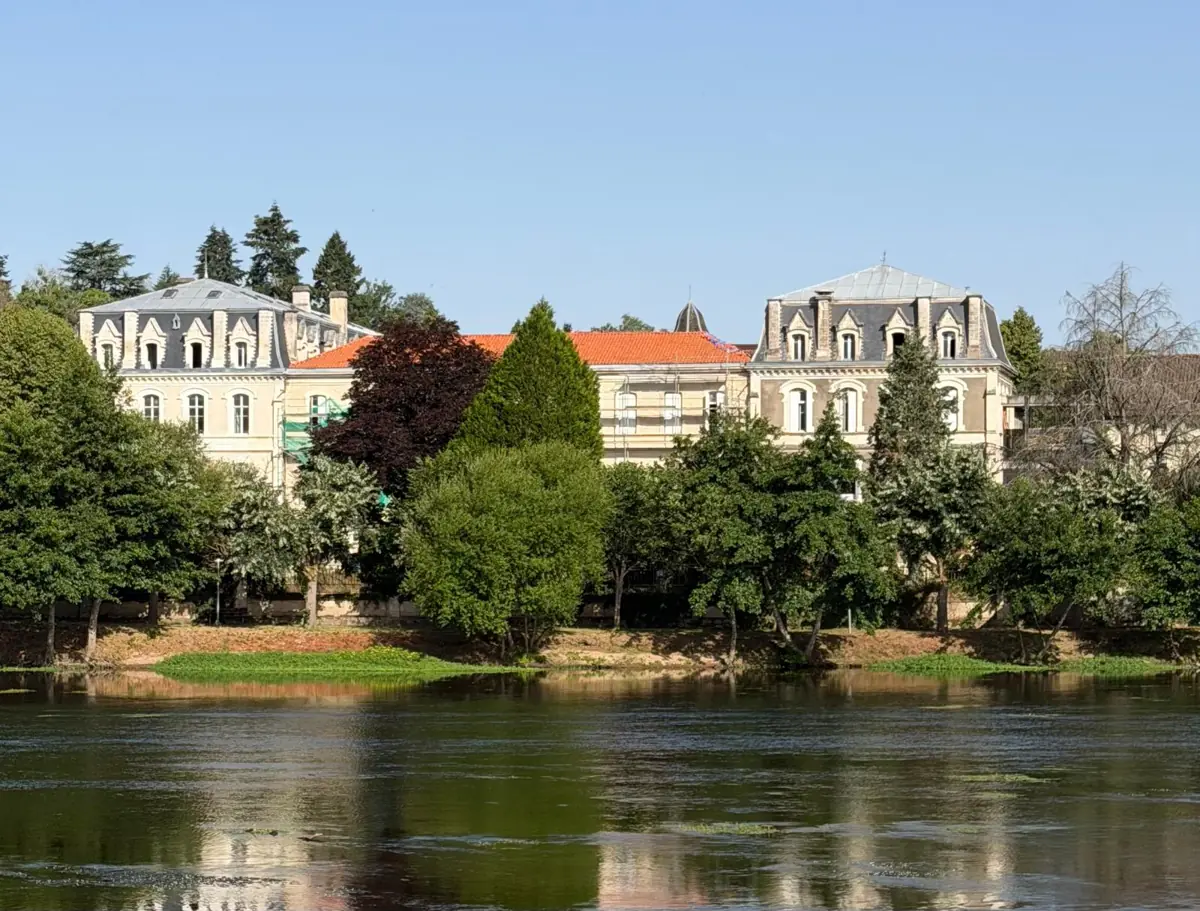 Grand pale stone buildings with slate mansard roofs behind a line of trees, reflected in a wide river