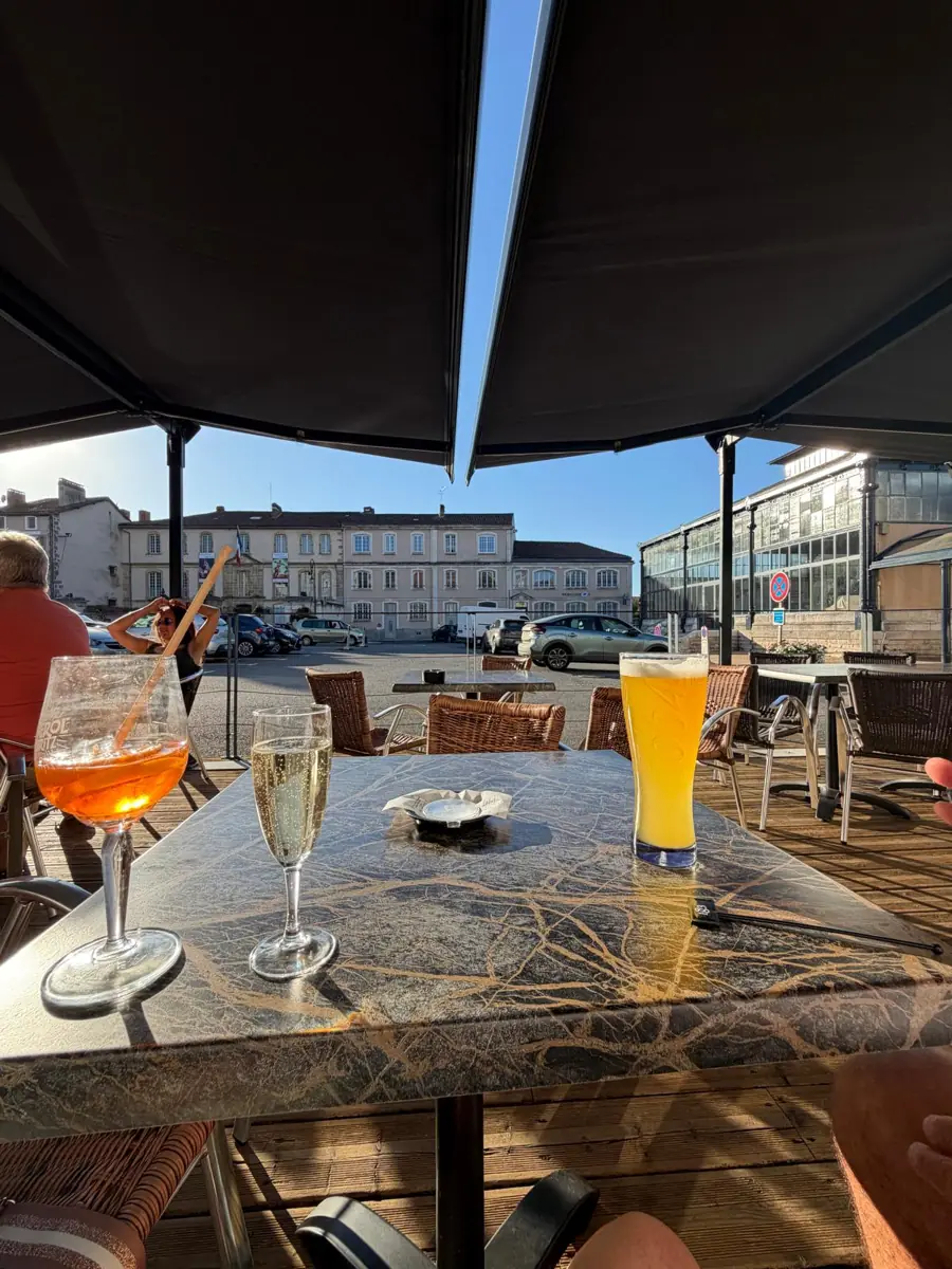 An Aperol spritz, a glass of fizz and a pint of beer on a marble-topped table under a dark parasol, looking out across a sunny square to tall pale town houses