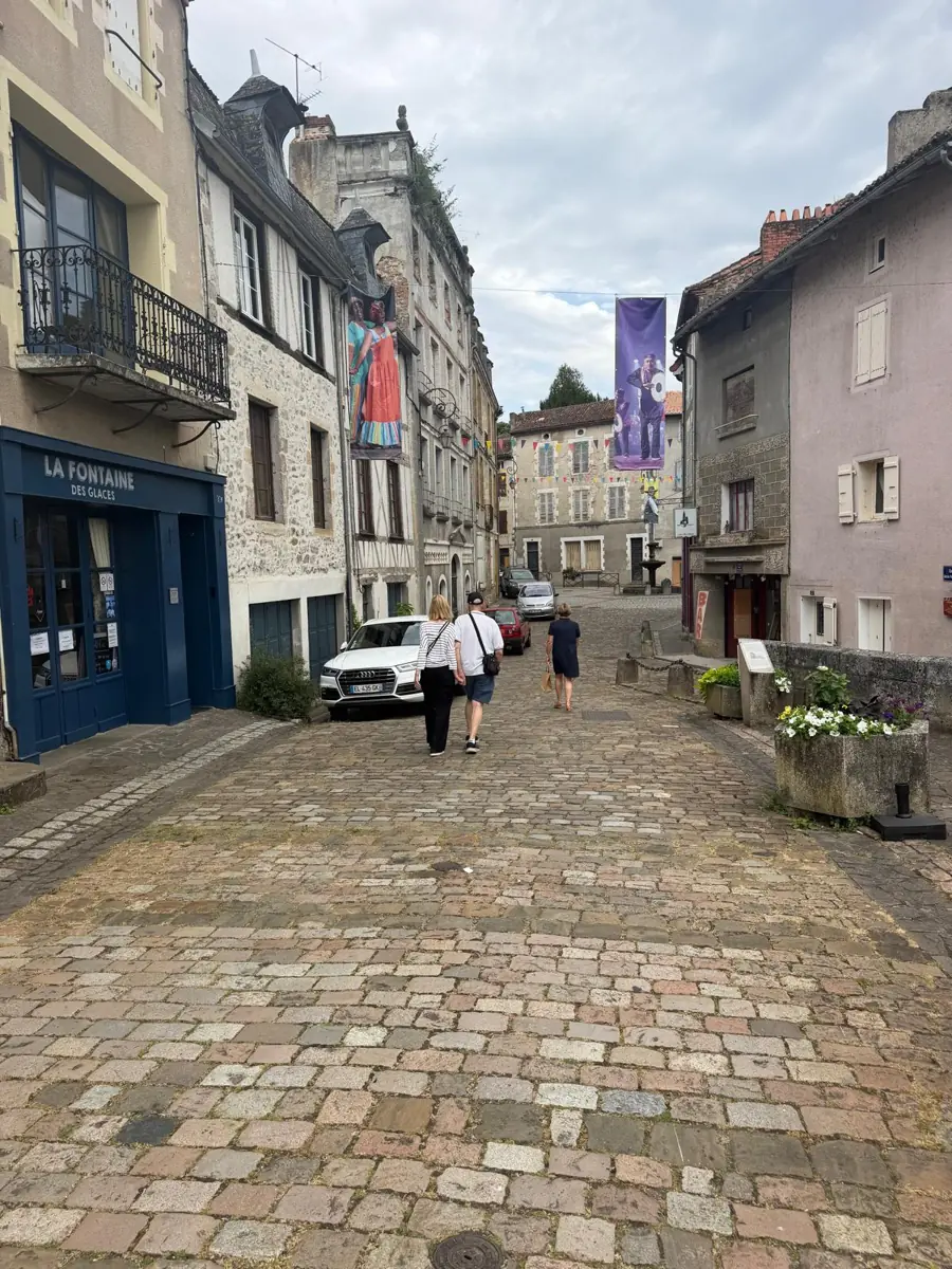 A wide cobbled street with shopfronts, balconies and a purple banner, people strolling towards a square