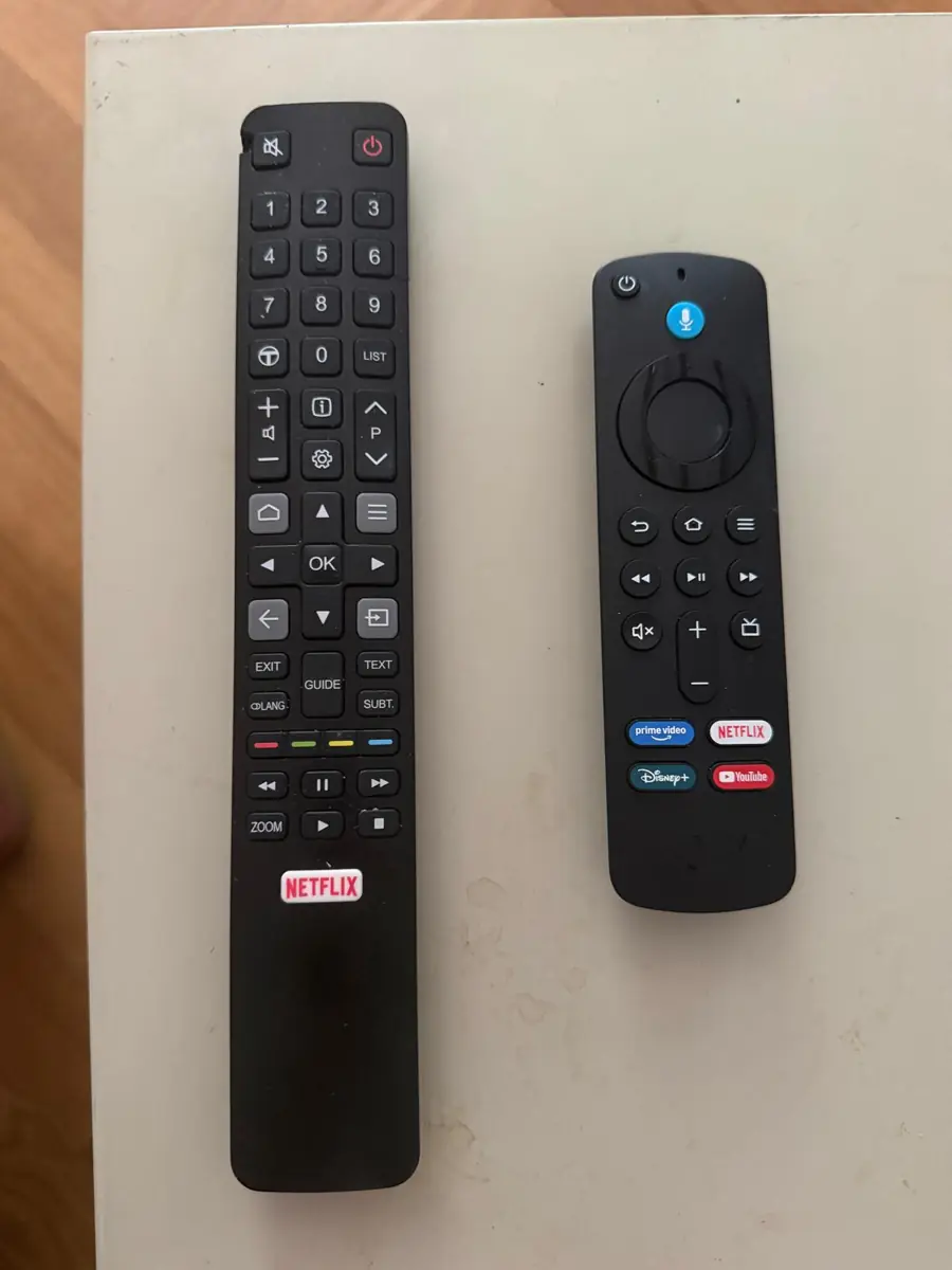 The two remotes side by side: the long black TV remote with a grey house-shaped Home button halfway down, and the shorter Amazon Fire Stick remote with Prime Video, Netflix, Disney+ and YouTube buttons
