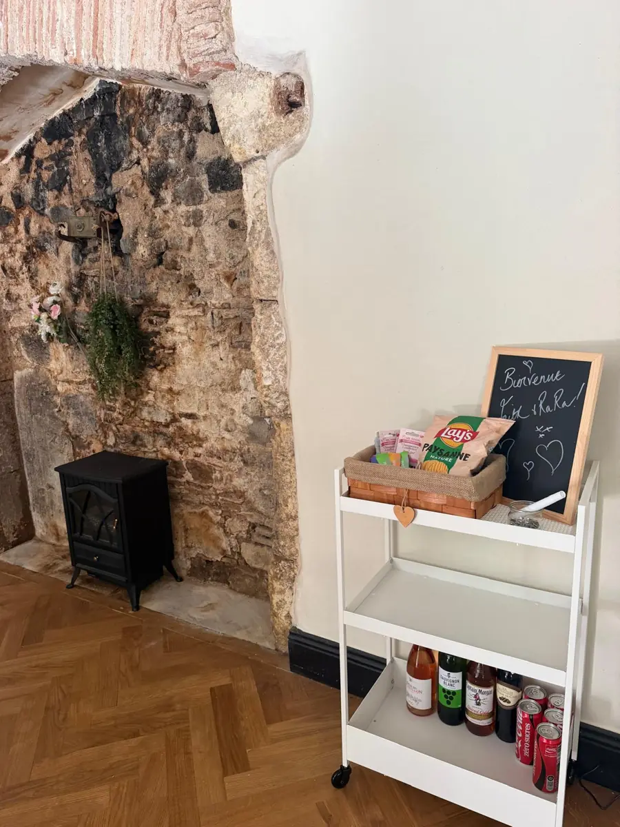 A white trolley against an old stone wall with a chalkboard reading Bienvenue, a basket of snacks and bottles of drink on the shelves below